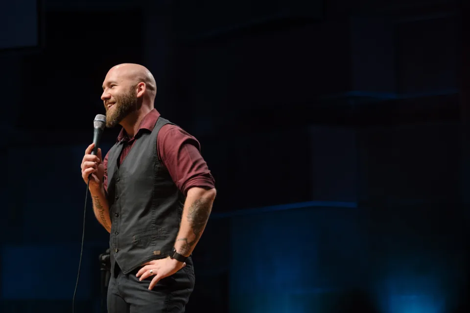 Tom smiling on stage, holding a microphone, wearing a burgundy shirt and dark waistcoat.