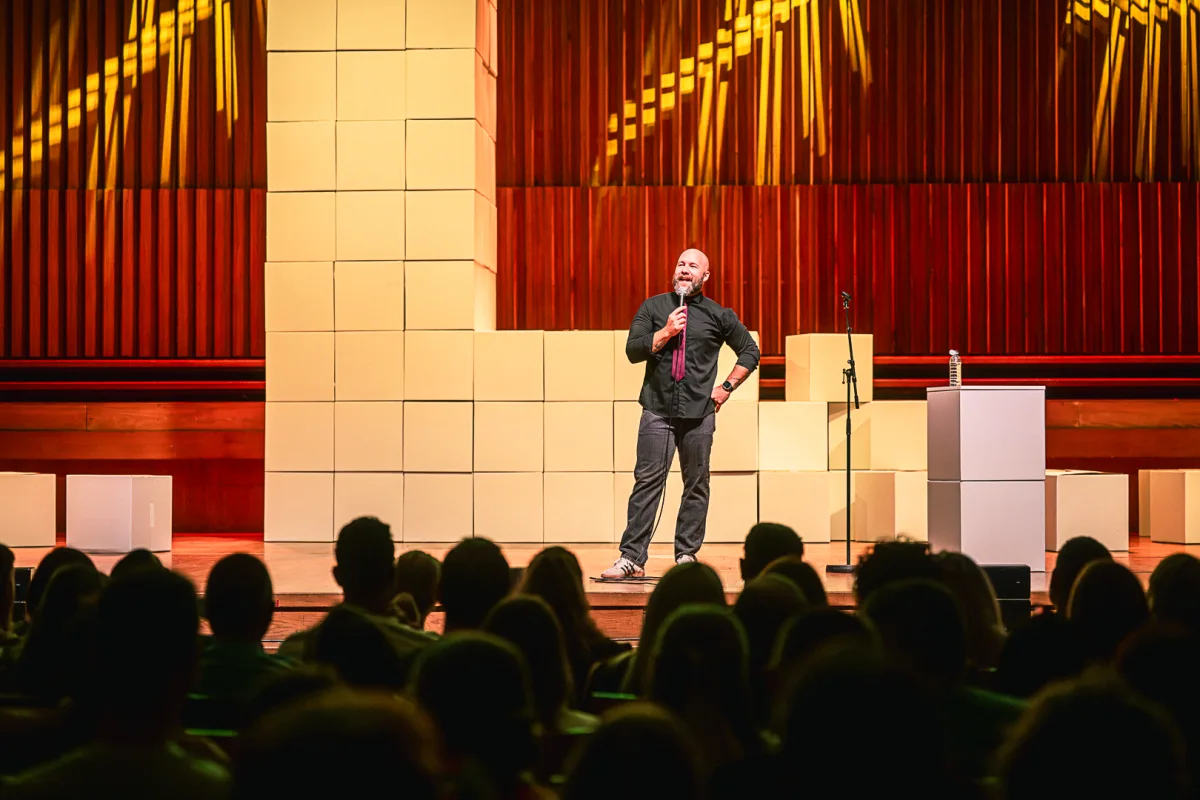 Tom performing stand-up at Lisinski, microphone in hand, with the audience in the foreground and a warmly lit stage behind him.